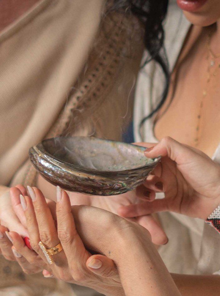 Mãos de Elini e de outra pessoa segurando uma concha com fumaça de ervas